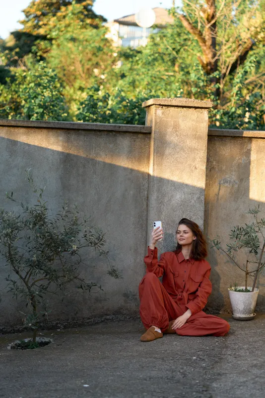 Woman in a red outfit sits on the ground in a sunlit courtyard, taking a selfie with a smartphone, conveying casual lifestyle and warm, relaxed self-expression