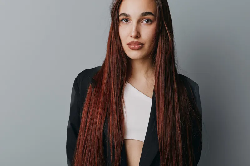 Young woman with long braided hair wearing black blazer and white top posing against gray background. Confident female model with natural makeup and stylish outfit for fashion or portrait photography.