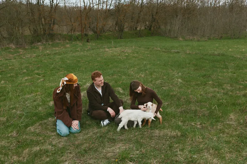Three people in a grassy field interact with a small white goat kid, enjoying a playful outdoors moment, creating a warm lifestyle scene ideal for family, nature, and animal projects.