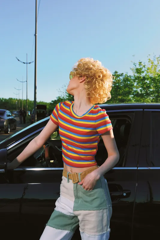Candid portrait of a young woman with curly blonde hair leaning against a black car on a sunny day, wearing a rainbow striped crop top and patchwork jeans, exuding carefree street style and