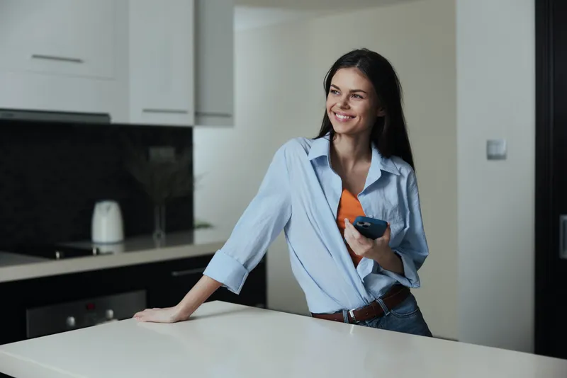 Женщина стоит на кухонной стойке, держит в руках мобильный телефон и смотрит в камеру в домашней обстановке