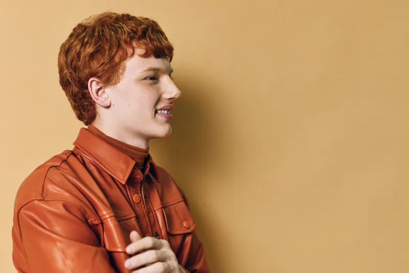 Young man with red hair smiling in orange leather jacket against plain beige background looking sideways with folded arms and relaxed confident posture in casual style.