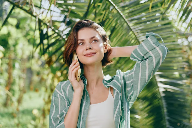 A cheerful woman stands outdoors among palm leaves, talking on a phone, wearing a striped green shirt and white top, with a relaxed, confident smile.