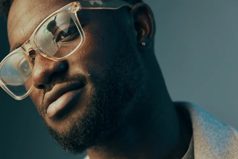 Portrait of a stylish Black man with glasses, showcasing confidence and modern fashion in a soft, cool toned background, ideal for themes of self expression and individuality