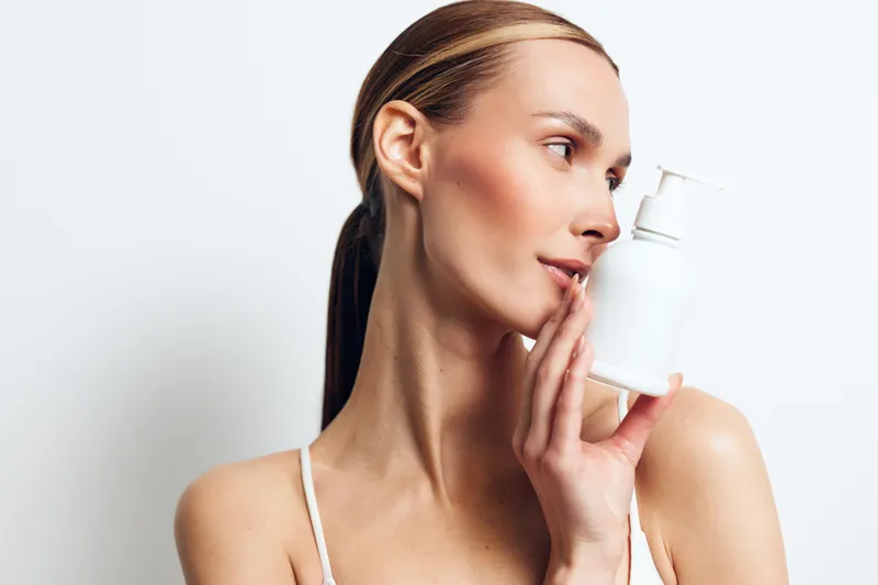 Young woman with clear skin holding white cosmetic bottle near face, minimal makeup, natural look, smooth complexion, soft lighting, clean background, beauty concept