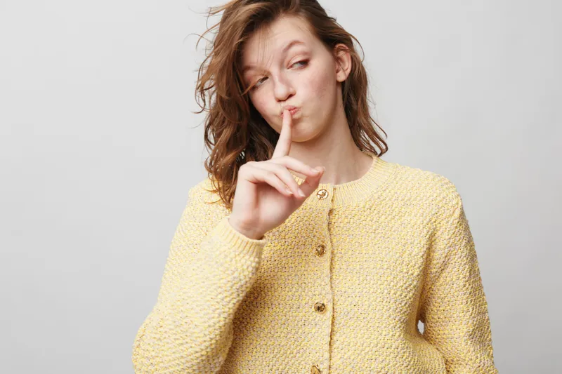Young woman in yellow sweater makes shushing gesture with finger on lips, eyes closed, isolated on white background. Concept of silence, secrecy, quiet, and calm expression.