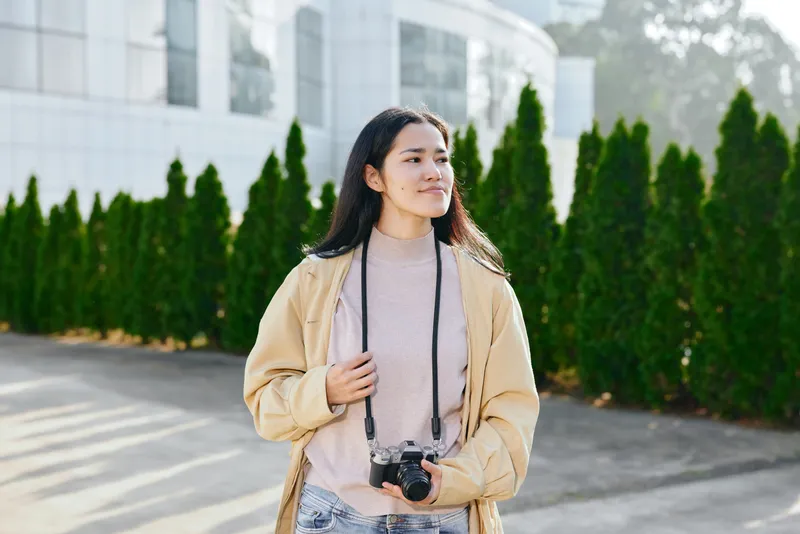 Young woman with a camera standing outdoors near a modern building, wearing a beige jacket and soft sweater, capturing a candid moment in daylight, emphasizing photography and everyday style