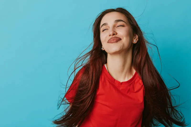 Young woman with long hair wearing red shirt standing against blue background with eyes closed and smiling expression. Casual portrait of happy female enjoying moment in studio.