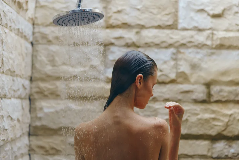 Woman showering outdoors under rain showerhead against natural stone wall, enjoying refreshing water spray in serene bathroom setting with modern rustic design.