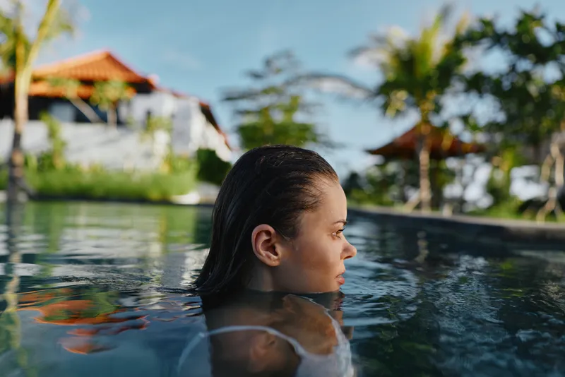 Young woman swims in outdoor pool in tropical resort with palm trees and traditional buildings in background enjoying summer lifestyle and relaxation.