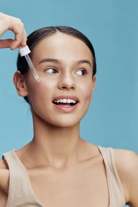 Young woman applying skincare serum with a dropper, showcasing a radiant smile against a light blue background, emphasizing beauty and self care