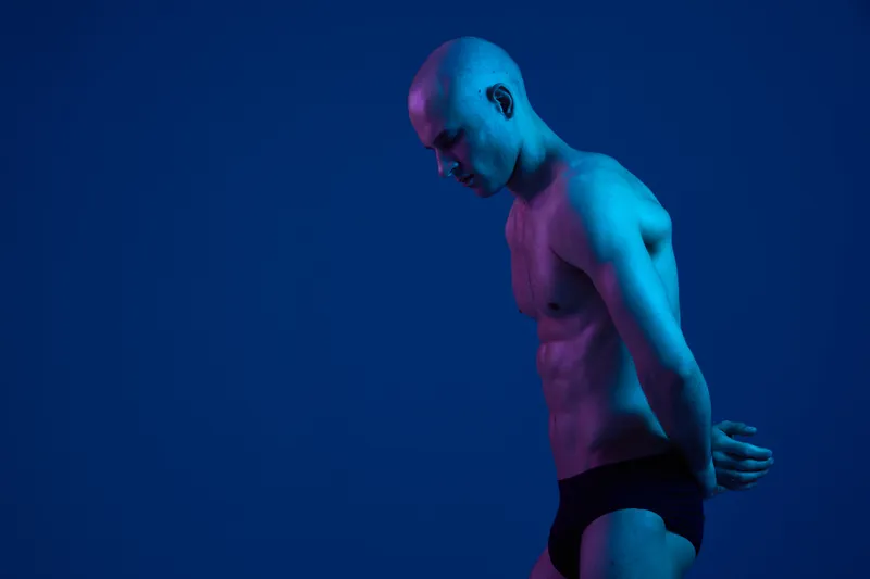 Man standing in a blue-lit studio, showcasing a strong physique with a contemplative expression. The image features stark contrasts and an artistic light play.