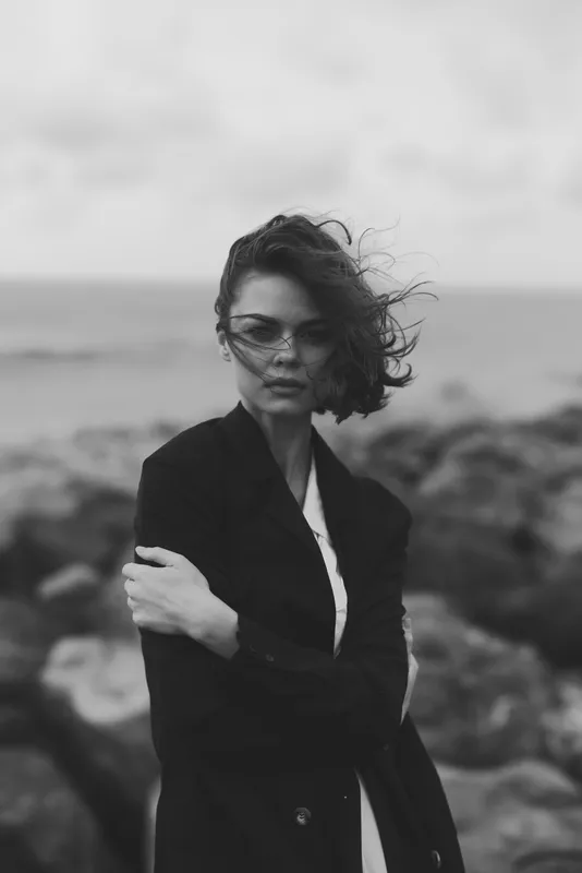 Moody monochrome coastal scene with a windblown coat and ocean breeze, showcasing a woman among rocks and shoreline to convey fashion, elegance and contemplative mood against gray skies