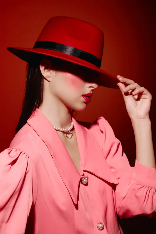 Profile of fashionable woman wearing bright red hat and coral pink suit with pearl necklace. Studio shot on red background inspired by creative beauty and fashion concepts.