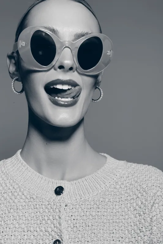 Young white woman with fair skin wearing large round sunglasses and earrings poses playfully with tongue out in a studio portrait. Fashion and fun concept.