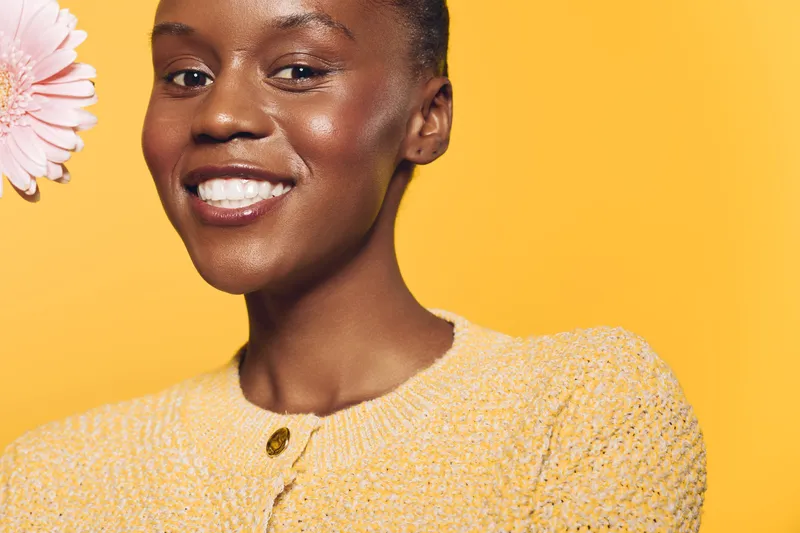 Smiling African American woman with dark skin wearing a yellow sweater poses against a yellow background holding a pink flower. Concept of beauty, happiness, and natural skincare.