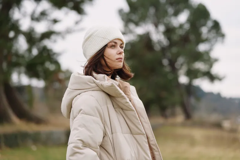 Winter woman in an oversized coat and beanie stands outdoors, calm and stylish against a natural landscape. Soft light highlights cozy mood and contemporary outdoor fashion.