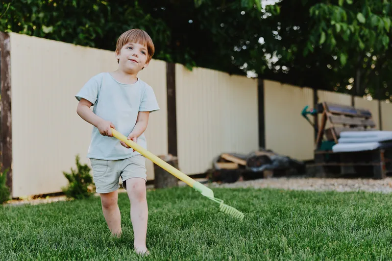 Happy boy playing outdoors with a yellow toy rake, enjoying a sunny day in a garden filled with greenery and laughter, promoting joy and playfulness.