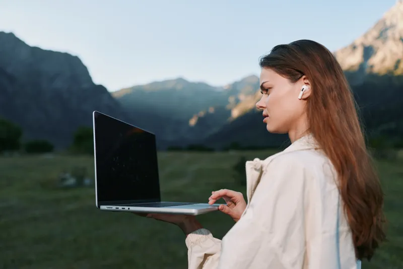 An attentive professional woman works on a laptop outdoors in a scenic valley. She wears wireless earphones, is calmly focused, and conveys productivity in a fresh, modern work setting.