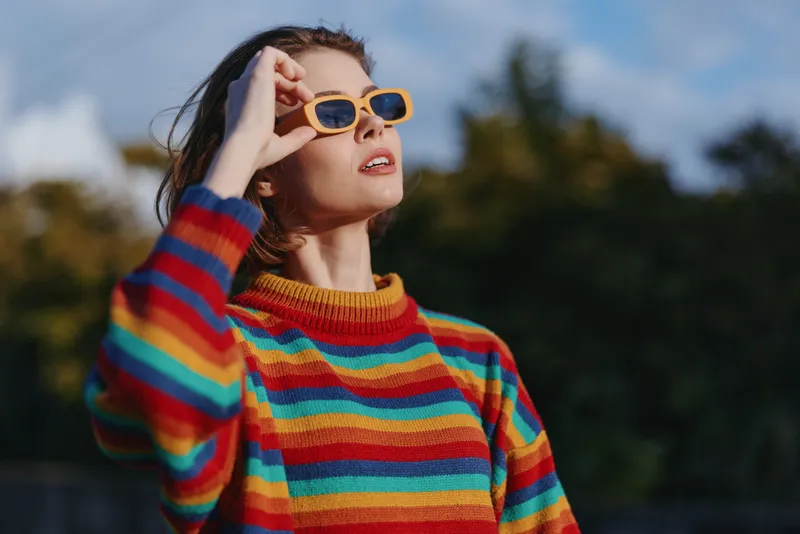 Woman in a striped sweater and yellow sunglasses portrait outdoor, looking up — young 20s model with shoulder-length brown hair, casual outfit, confident travel lifestyle fashion vibe.