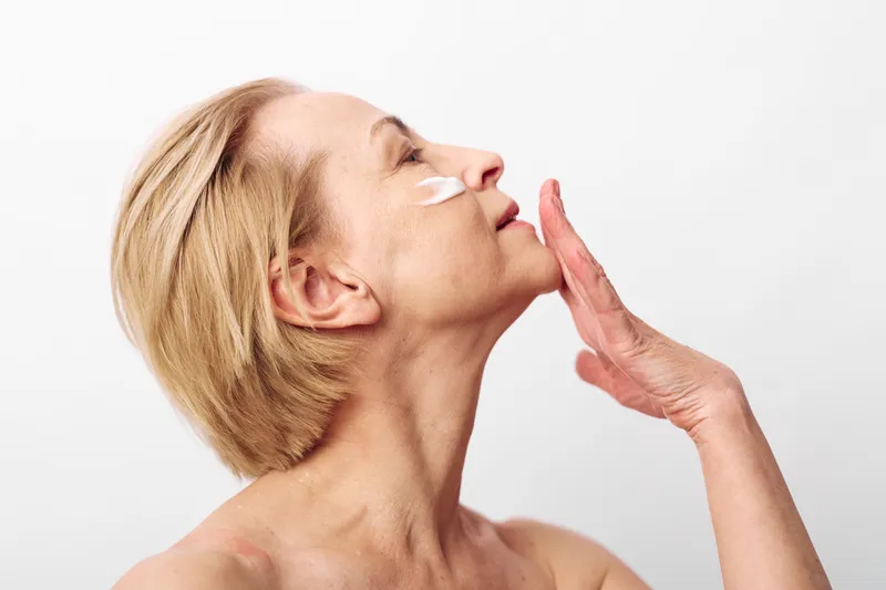 Mature woman with short blonde hair applies white face cream on cheek in bright studio. Concept of skincare, aging skin care, beauty routine, and self-care for mature women.