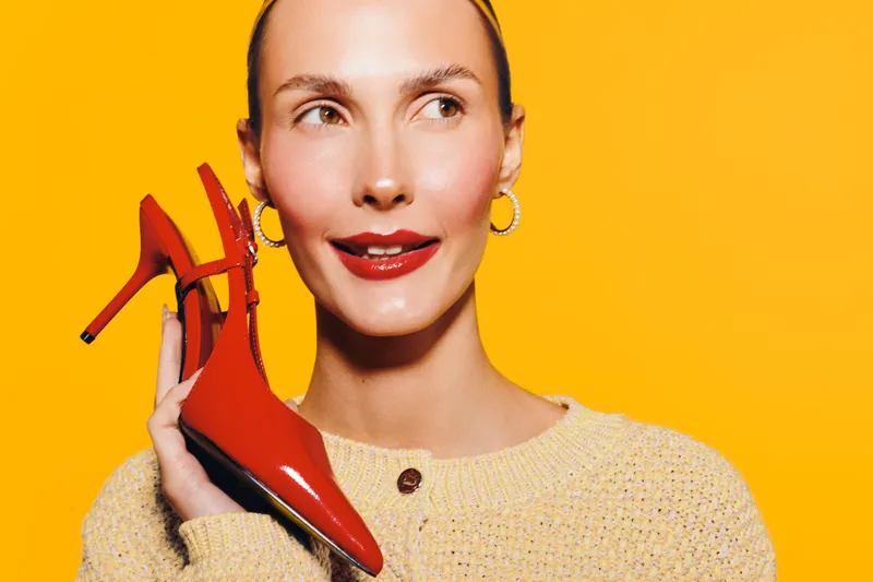 Smiling woman with red high heel shoes near her face on bright yellow background. Fashion concept with stylish footwear and joyful female model wearing beige sweater and hoop earrings.