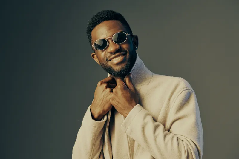 Stylish young man with sunglasses and a fashionable beige coat, smiling confidently against a simple green background
