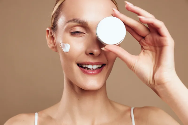 Smiling woman with clear skin holding white cream jar near eye, applying face moisturizer, natural beauty, skincare routine, beige background, healthy glowing complexion
