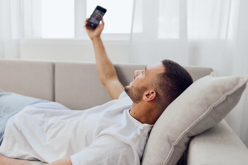 Smiling man relaxing on a couch while using a smartphone, enjoying a casual moment at home with natural light streaming in. Perfect for lifestyle themes.
