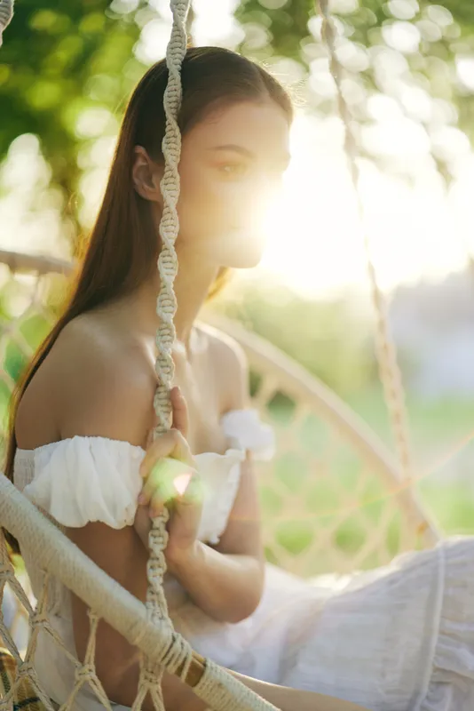 Young woman in white dress sitting on a swing, illuminated by sun rays, exuding tranquility and joy in a lush green garden, conveying a sense of serenity and connection to nature