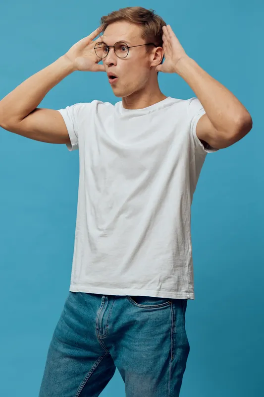 surprised young caucasian man wear white t-shirt and glasses hold hands near head stand against blue background digital detox concept