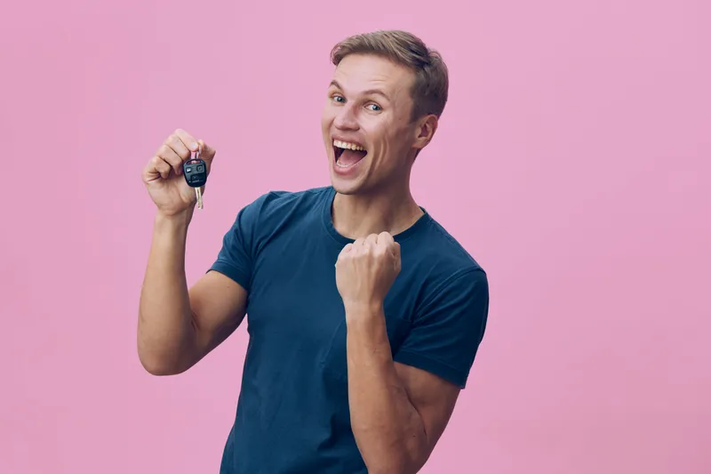 Smiling man holding car key with excited expression against pink background. Happy, joyful, casual, young adult, celebration, lifestyle concept.