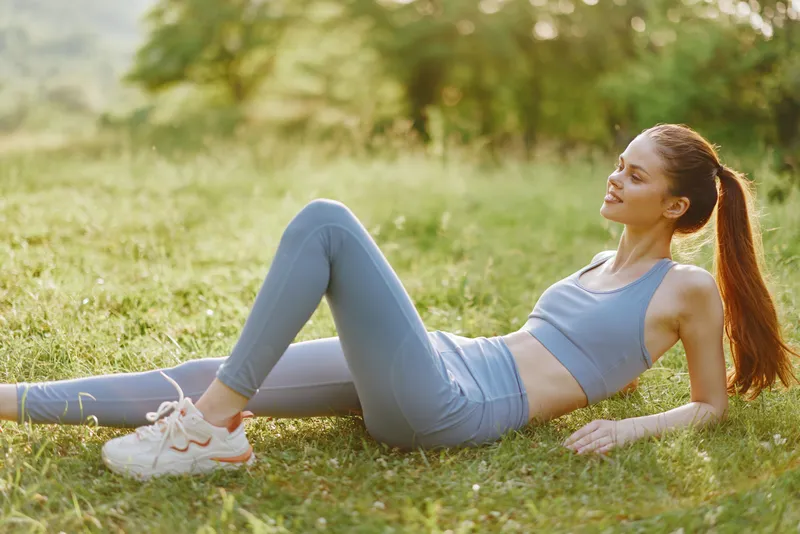 Young woman in activewear enjoying a peaceful moment in nature, demonstrating relaxation and fitness in a green outdoor setting with a bright color palette
