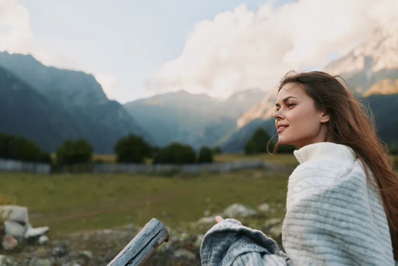 Smiling woman in a white knit sweater enjoying a scenic outdoor setting with mountains, fields, and a clear sky, conveying lifestyle, relaxation, and nature appreciation.