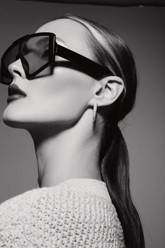 Profile of a stylish Caucasian woman with fair skin wearing large sunglasses and hoop earrings. She has slicked-back hair and a textured sweater in a studio setting.
