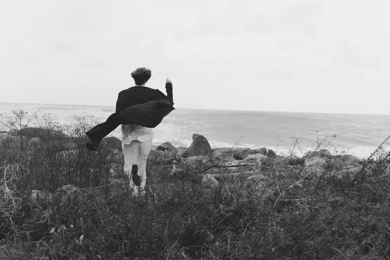 Silhouette scene of two figures on a windy coast, one lifting the other, exploring rugged terrain and sea horizon, conveying freedom, adventure and bold travel moment in monochrome tones