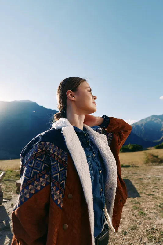 Woman in mountains wearing a warm jacket outdoors, portrait in nature with sunlight on face and meadow background, young traveler with braided hair enjoying scenic landscape.