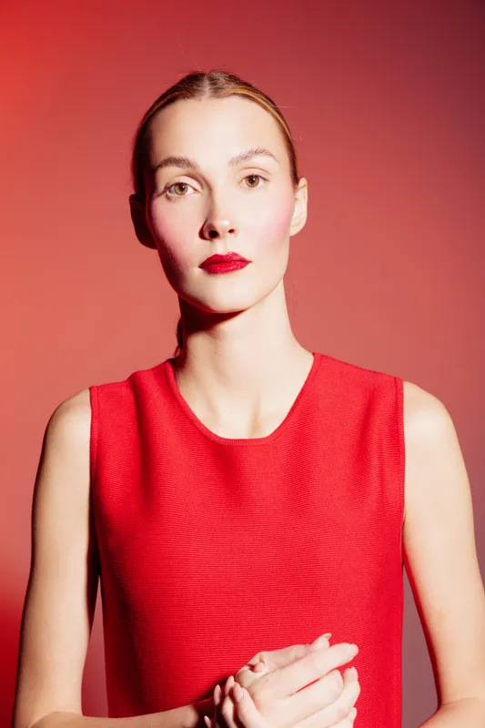 Young woman with red lipstick and sleeveless red dress poses against red background. Studio portrait with strong lighting highlights facial features and confident expression.