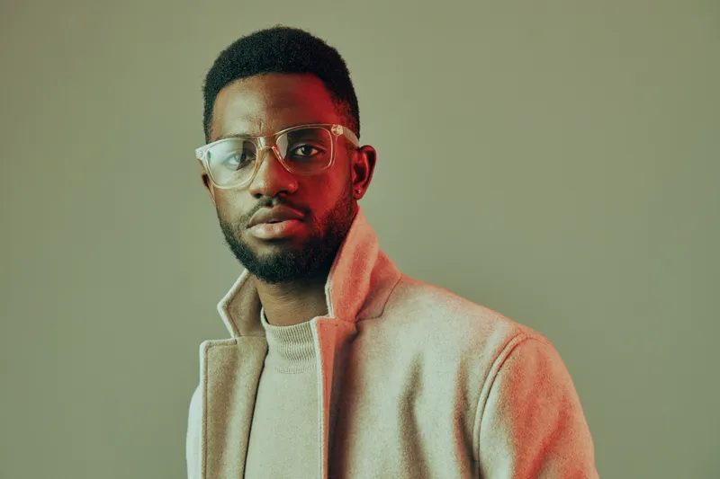 Stylish young man in a beige coat with glasses posing against a soft green background, exuding confidence and sophistication, perfect for fashion and lifestyle concepts