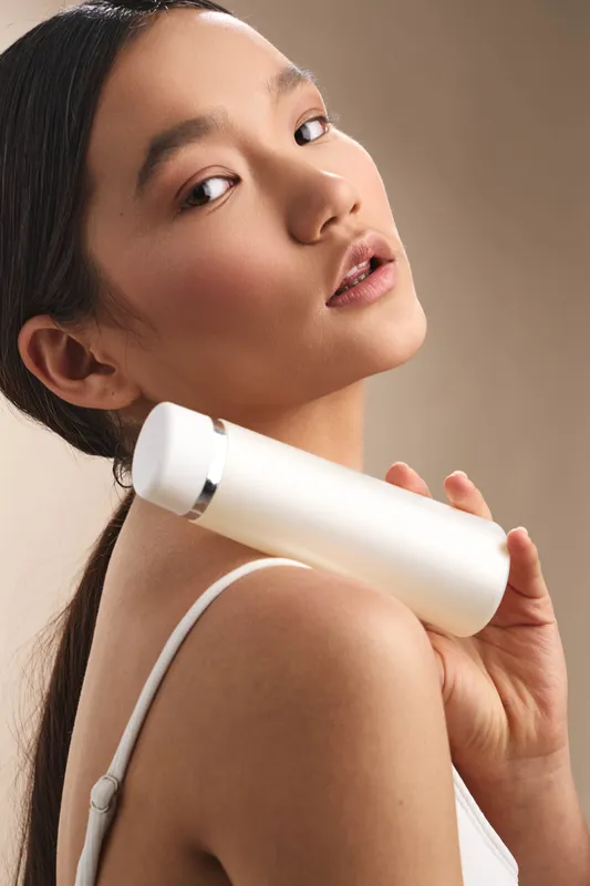 asian woman holds a white cosmetic bottle close to her shoulder in a soft studio setting. she looks toward the camera with a gentle smile, conveying skincare focus, calm confidence, and modern beauty