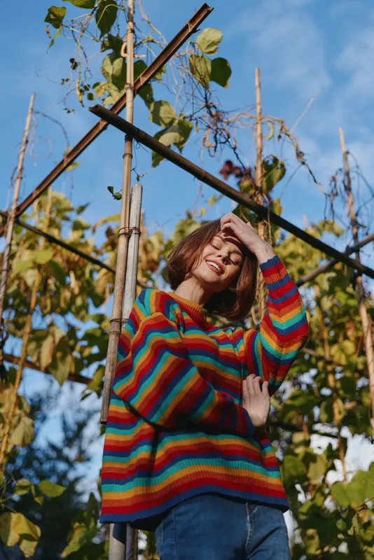 Young woman, rainbow sweater, trellis, garden: smiling young woman in a colorful striped sweater leaning on a wooden trellis in a sunlit garden, casual jeans, short bob hair, joyful outdoor lifestyle
