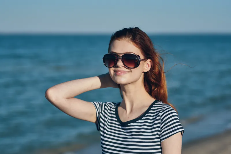 Женщина в солнечных очках стоит на пляже у моря, на заднем плане — природный пейзаж