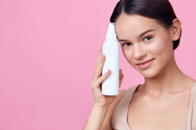 Young woman applying hair care product with confidence, soft smiles, pink background, promoting healthy hair and self care routine