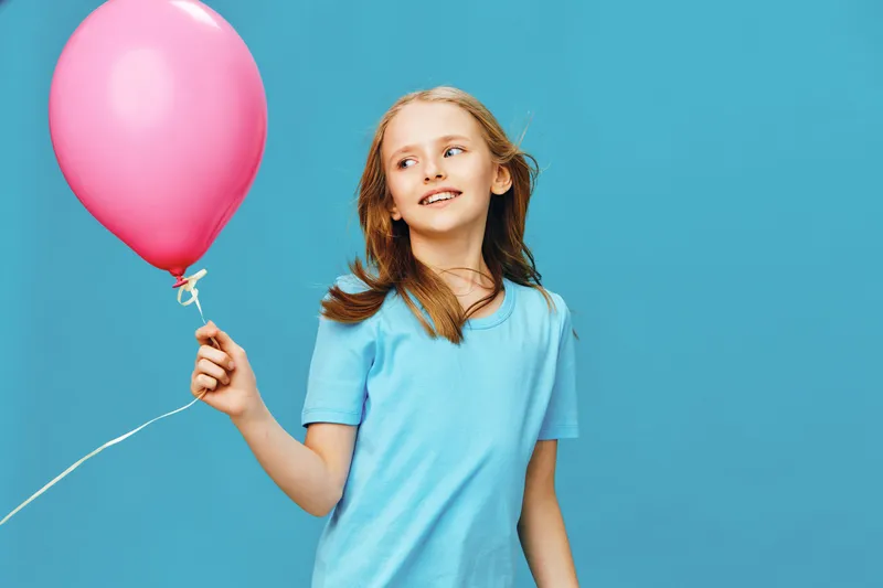 Joyful young girl holding a pink balloon against a vibrant blue backdrop, showcasing happiness and playfulness in a cheerful, bright atmosphere