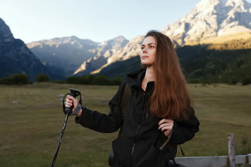 A young woman stands in a rugged mountain landscape, wearing a black jacket and gripping a trekking pole as she gazes into the distance, ready for outdoor adventure.