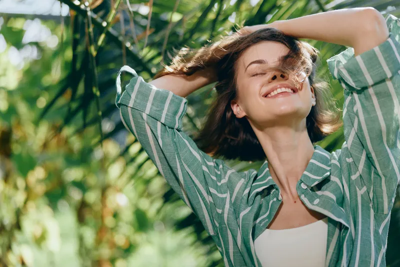 Woman outdoors, smiling under bright sunshine, surrounded by greenery. A relaxed pose in casual clothing conveys joy, freedom, and a fresh breeze on a warm day in nature.