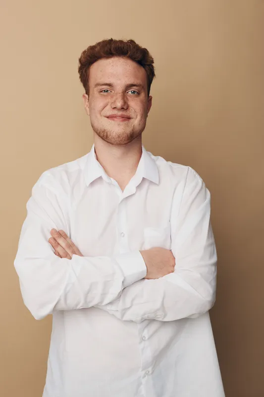Confident young man with curly hair wearing white shirt stands with crossed arms against beige background. Portrait of casual male model with slight smile and relaxed posture in studio.