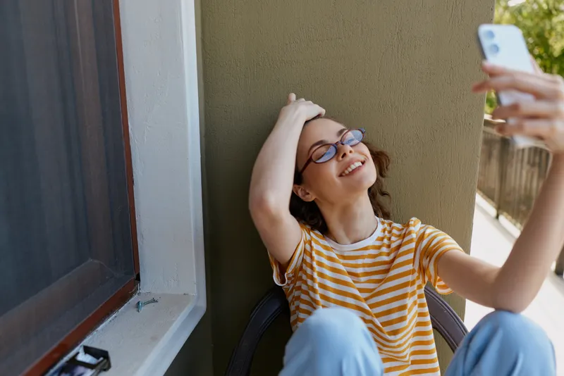 Young woman with brown hair and glasses wears a striped shirt while posing for a casual selfie against a sunlit balcony wall, expressing a relaxed mood and everyday lifestyle.