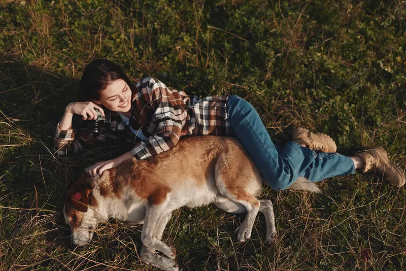 Woman relaxing outdoors with a dog on a grassy field, lying down and enjoying nature, wearing a plaid shirt and jeans, a peaceful moment in the open landscape