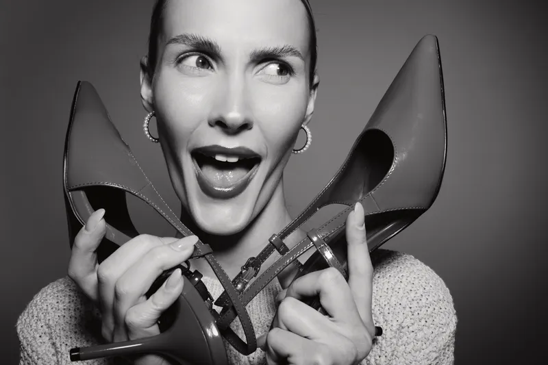 White woman with light skin holds pair of high heel shoes near face and looks surprised. Black and white portrait with expressive facial expression and fashion concept.